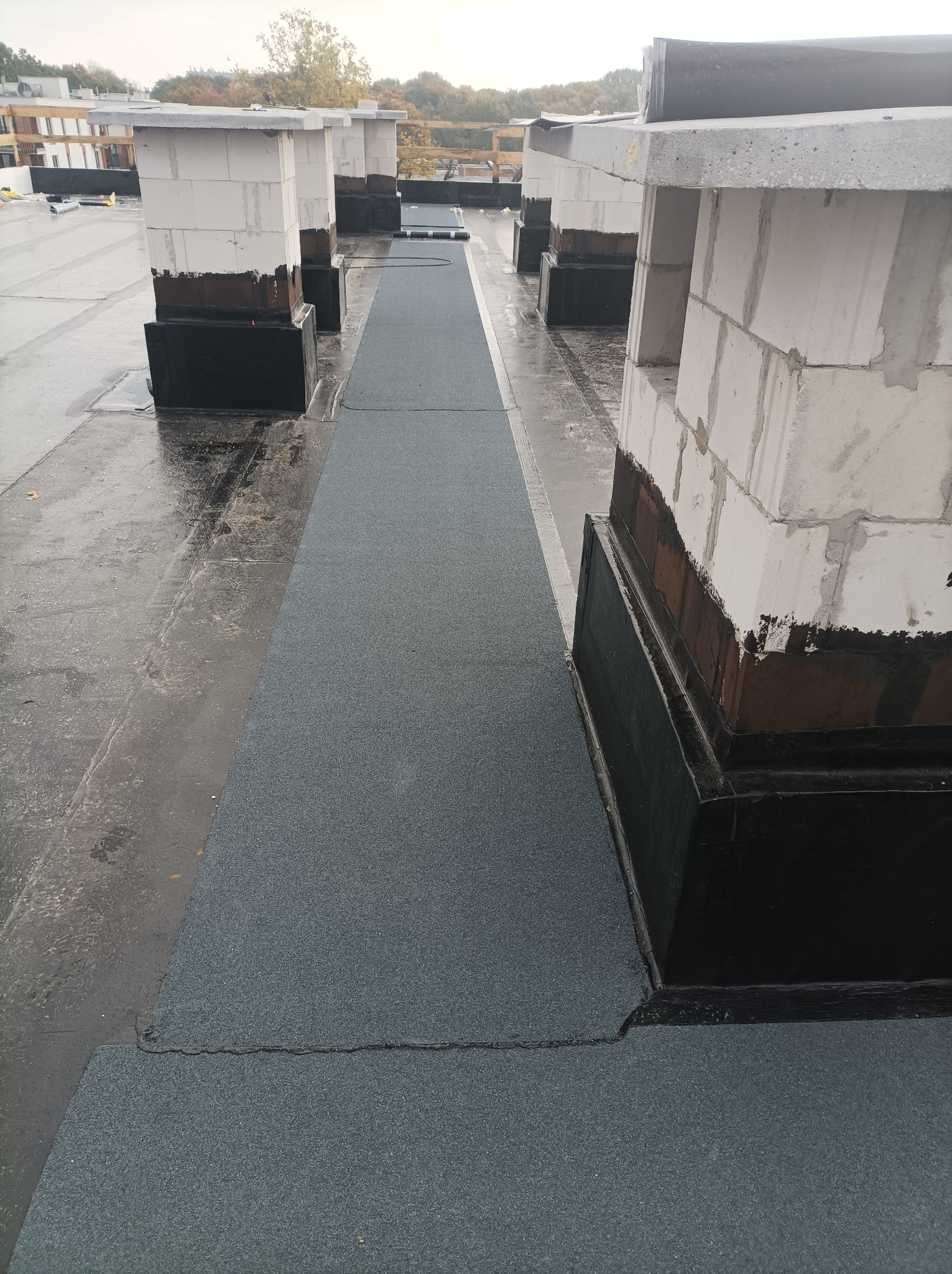 Szary pas papy termozgrzewalnej na płaskim dachu, widoczne kominy wentylacyjne z białych bloczków betonowych i obróbki blacharskie, lekko wilgotna powierzchnia dachu.