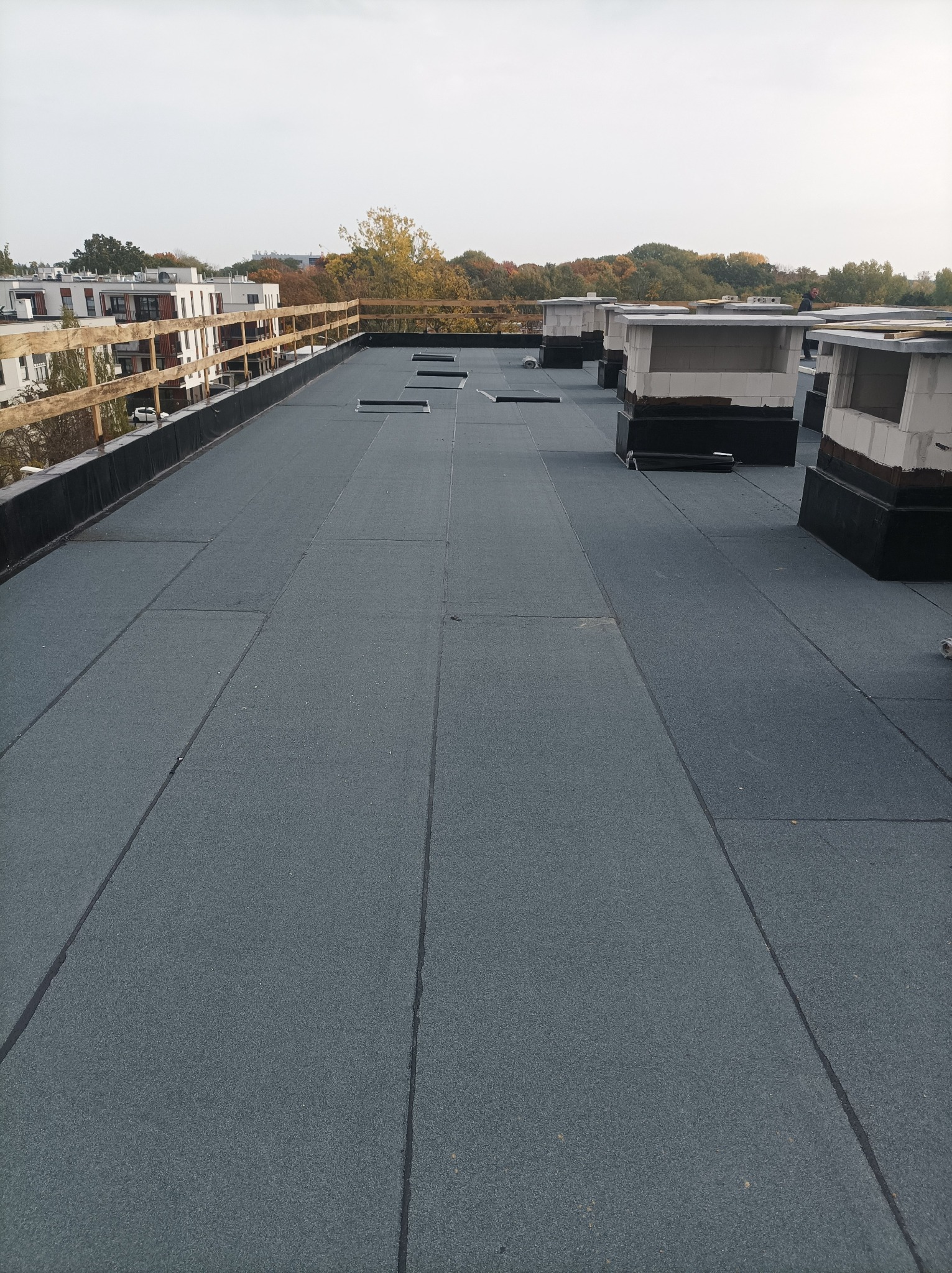 Płaski dach pokryty szarą papą termozgrzewalną, widoczne kominki wentylacyjne i drewniana balustrada na krawędzi dachu, w tle zabudowa miejska i drzewa.