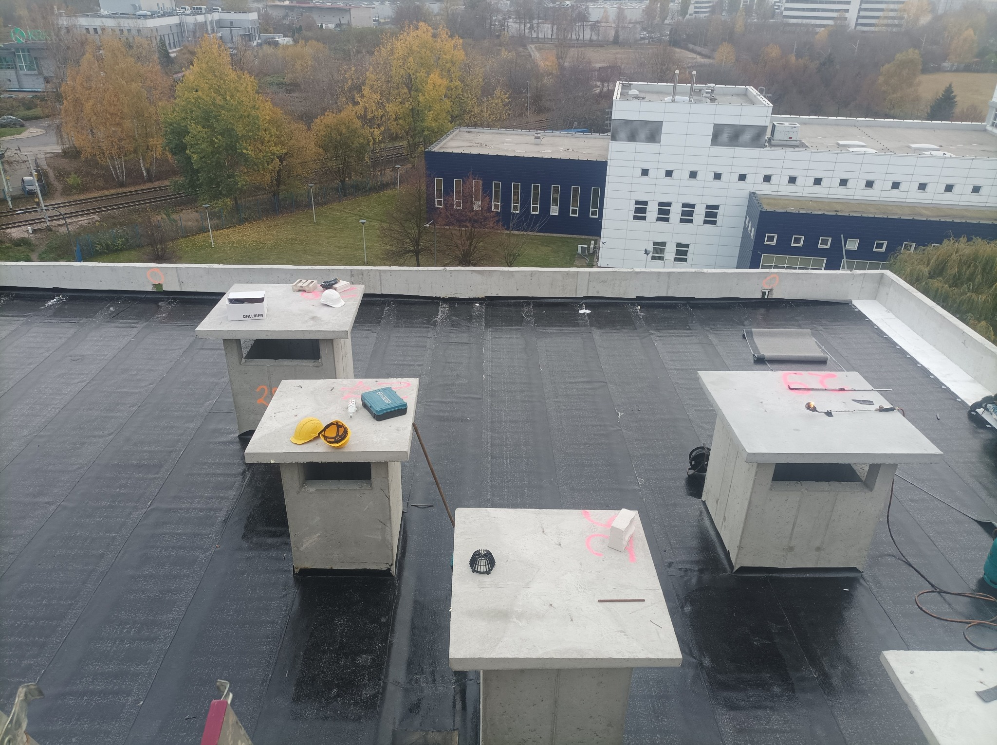 Widok z góry na płaski dach pokryty czarną papą termozgrzewalną, z czterema betonowymi podstawami wentylacyjnymi, na których leżą narzędzia, żółty kask i pudełko, w tle budynki i drzewa w jesiennej...