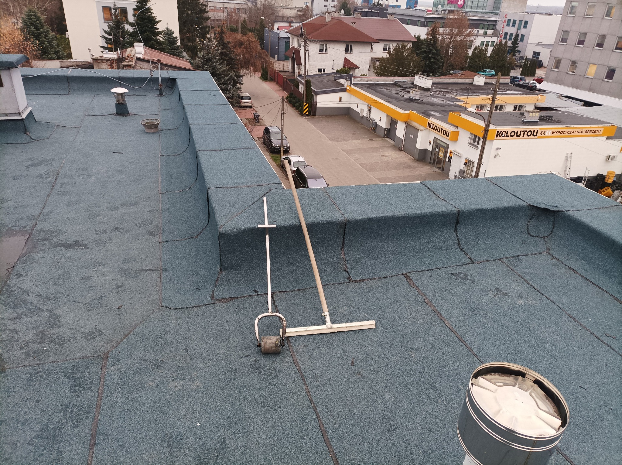 Widok z góry na płaski dach pokryty papą termozgrzewalną z widocznymi narzędziami do jej układania: wałek dociskowy i rozprowadzacz masy, w tle zabudowa miejska.