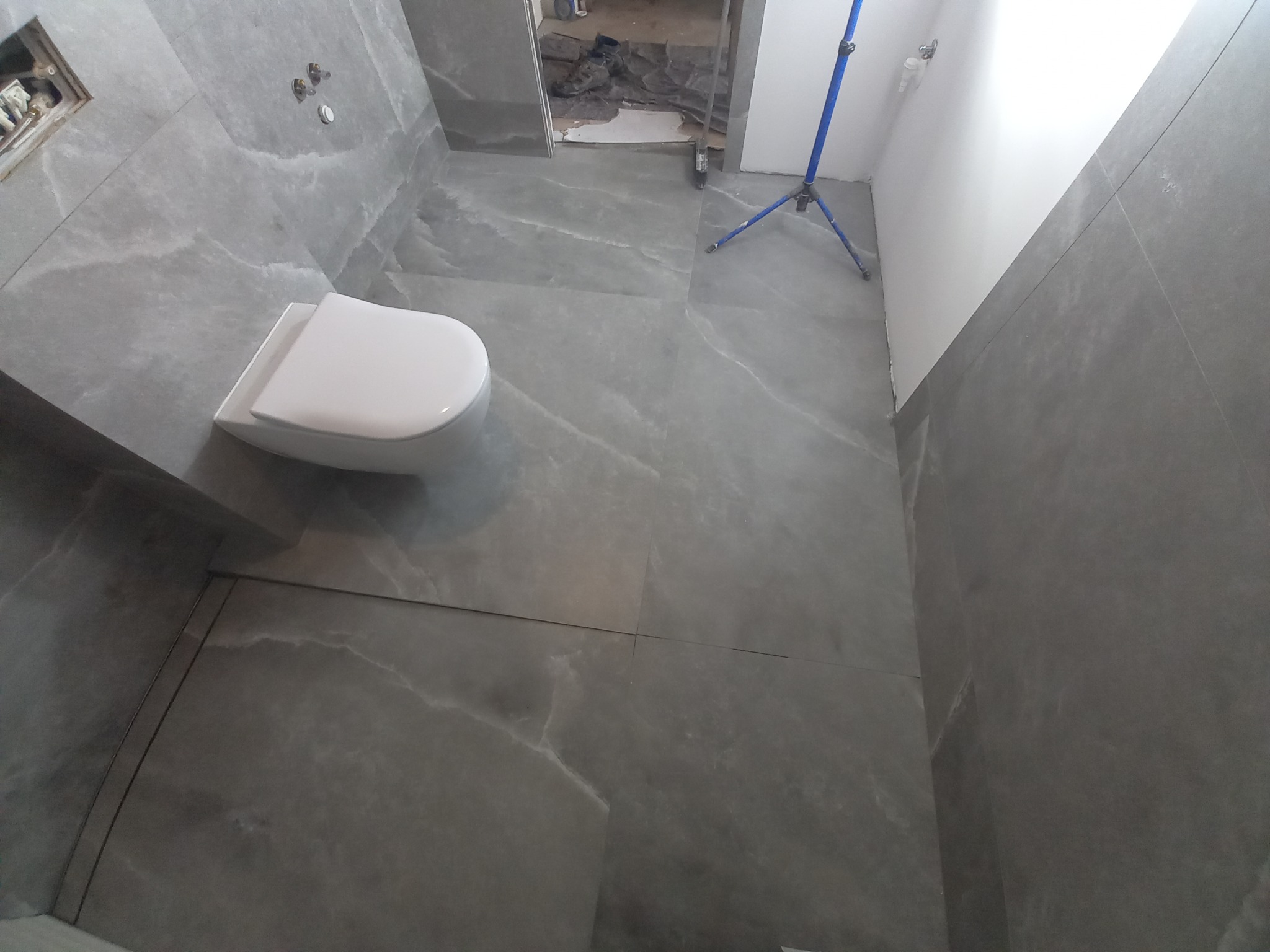 Szare płytki imitujące marmur na ścianach i podłodze w łazience w trakcie remontu, widoczna biała toaleta i niezagospodarowana przestrzeń za drzwiami.