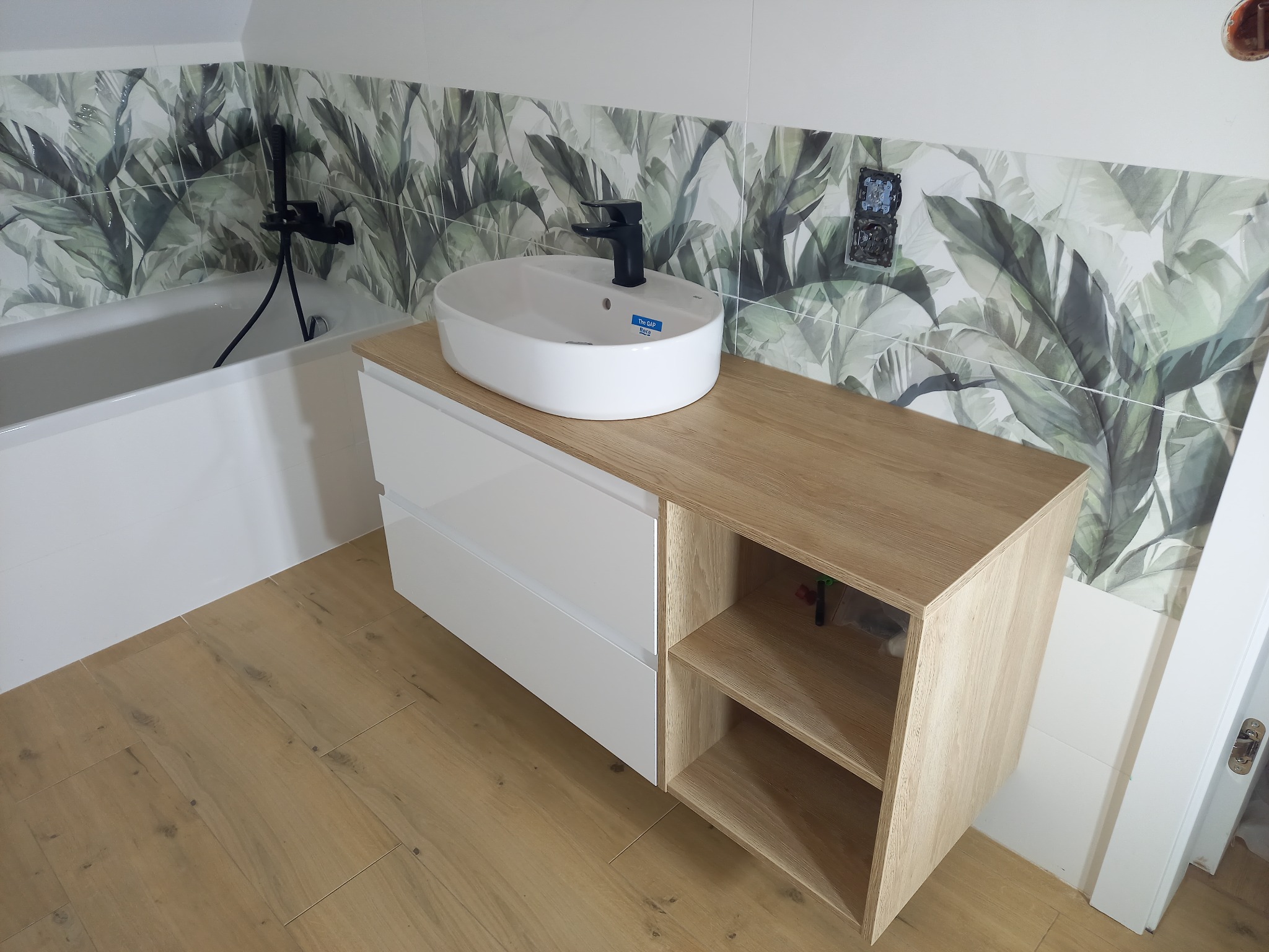 Nowoczesna łazienka z umywalką nablatową, szafką z drewnianym blatem i białymi frontami, oraz płytkami ściennymi w motywie tropikalnych liści.