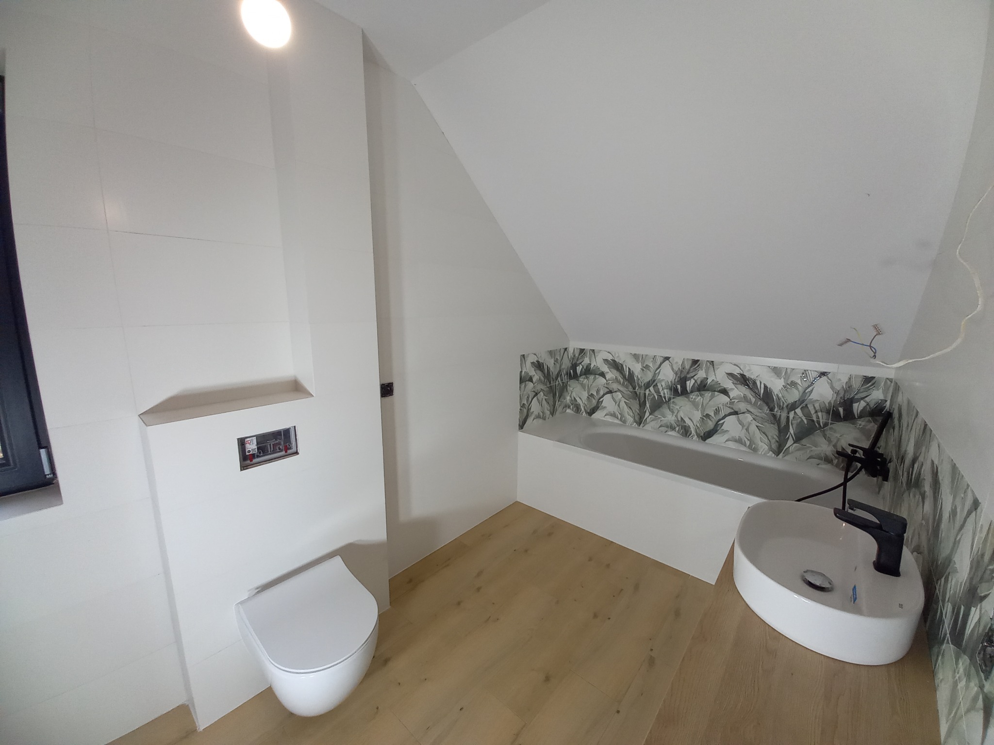 Nowoczesna łazienka na poddaszu z białymi płytkami, drewnopodobną podłogą, wiszącą toaletą, umywalką nablatową i wanną obudowaną płytkami z motywem roślinnym.