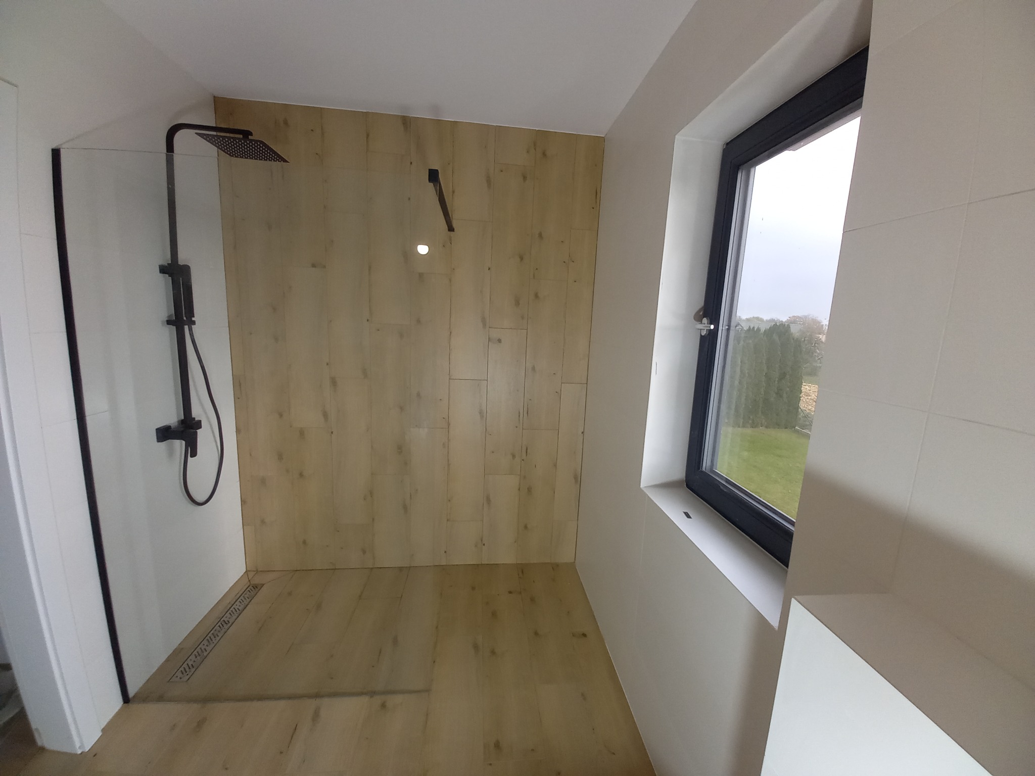 Nowoczesna kabina prysznicowa typu walk-in z czarną armaturą i odpływem liniowym, wykończona panelami imitującymi drewno, widok na zewnątrz przez okno.