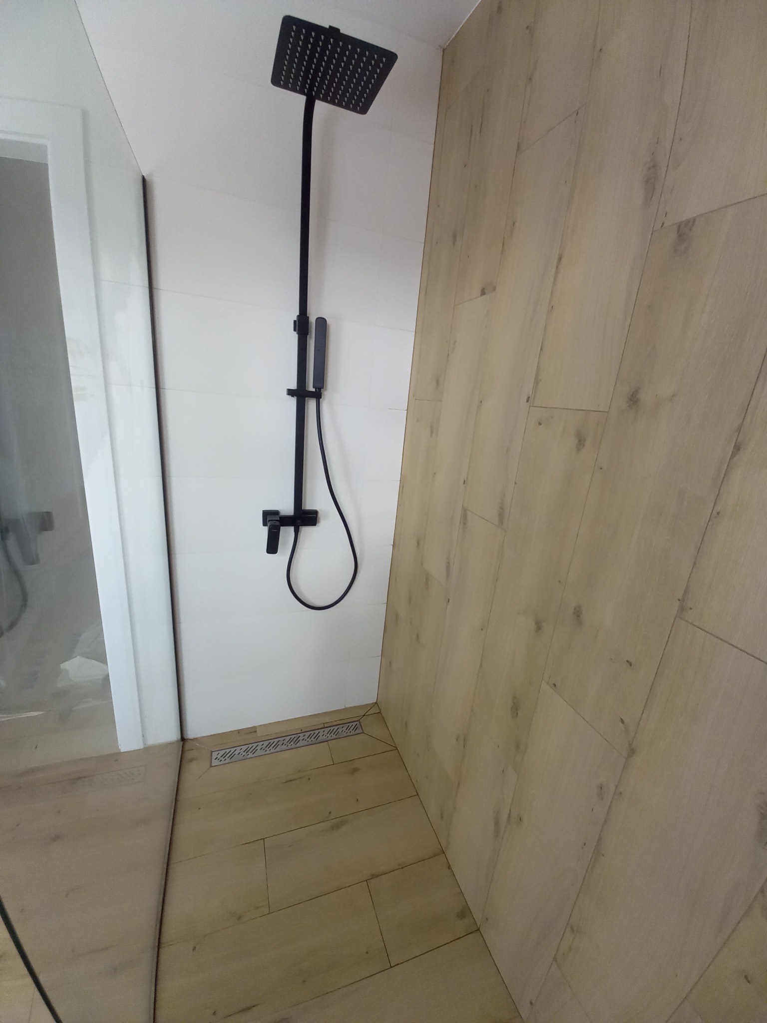 Nowoczesna kabina prysznicowa z czarną armaturą i odpływem liniowym, wykończona płytkami imitującymi drewno.