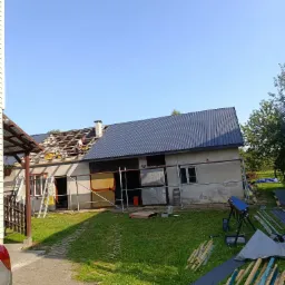 Dwupoziomowy budynek z częściowo wymienionym poszyciem dachowym. Na górze, pracownik w białej koszulce, z elektronarzędziem w dłoni. Część dachu pokryta szarą blachą trapezową, a druga część...