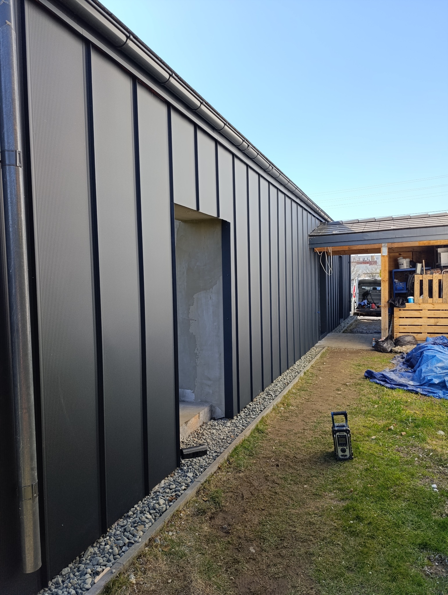 Nowoczesna elewacja budynku pokryta panelami w kolorze grafitowym, widoczne rynny i fragment podbitki dachowej, w tle fragment podwórka z narzędziem pomiarowym.