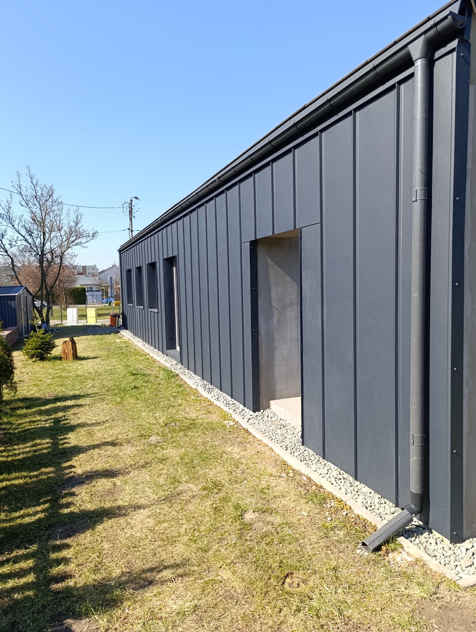 Nowoczesny budynek z szarą elewacją panelową, widoczne okna, niezadaszone wejście z surowego betonu, rynna spustowa i trawnik z elementami krajobrazu w tle.