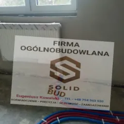 Tabliczka reklamowa firmy ogólnobudowlanej Solid Bud z logo, danymi kontaktowymi i hasłem reklamowym, ustawiona w surowym wnętrzu budowy z widocznymi rurami instalacyjnymi i grzejnikiem w folii.