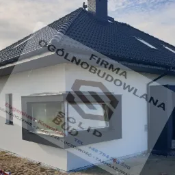 Biały dom jednorodzinny z ciemnoszarą stolarką okienną i dachem z dachówki ceramicznej, widoczny z boku, z elementami wykończeniowymi elewacji w trakcie prac, na pierwszym planie piasek i materiały...