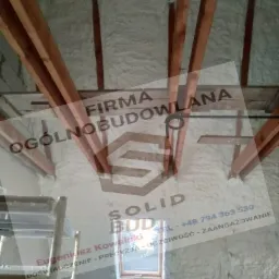 Poddasze z drewnianą konstrukcją więźby dachowej, zaizolowane pianą poliuretanową, widoczne okno dachowe i fragment rusztowania, na pierwszym planie półprzezroczyste logo firmy budowlanej.