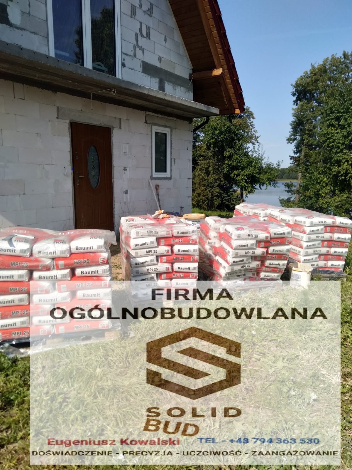 Niedokończony dom z białego bloczka, z oknem i brązowymi drzwiami, obok sterty zapakowanych materiałów budowlanych Baumit MPI 25, w tle drzewa i jezioro, na pierwszym planie logo firmy budowlanej...