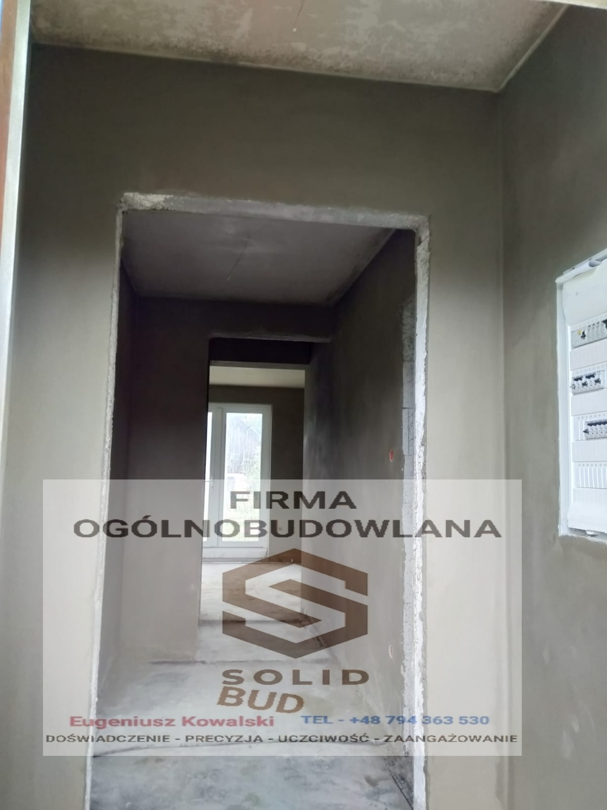 Korytarz w stanie surowym z widocznymi świeżo nałożonymi tynkami, przygotowany do dalszych prac wykończeniowych; w tle widoczne okno.