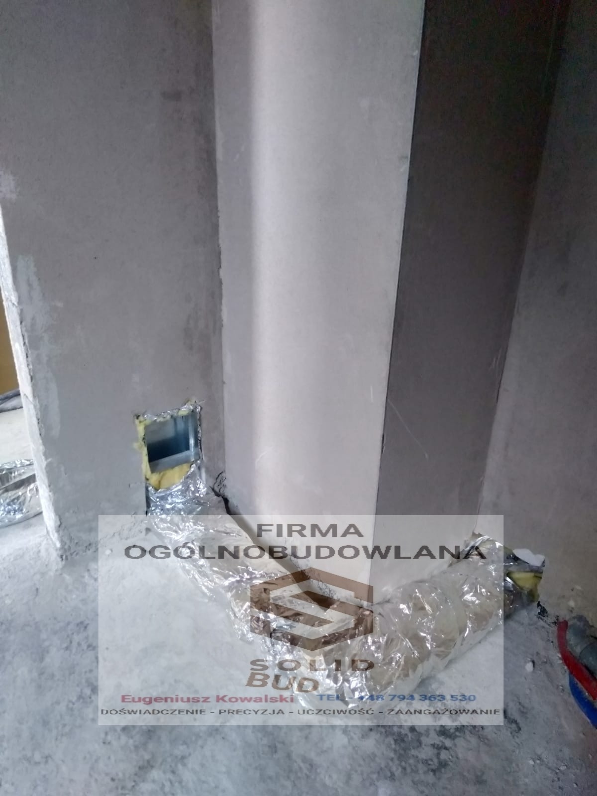 Niedokończone wnętrze z widocznymi przewodami zabezpieczonymi folią aluminiową i otworem w ścianie wypełnionym żółtą izolacją, na pierwszym planie folia ochronna z nadrukiem nazwy firmy budowlanej.