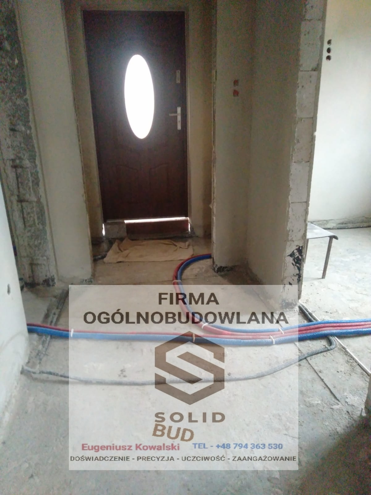 Koszalin: Wnętrze w trakcie remontu z widocznymi instalacjami hydraulicznymi i drzwiami wejściowymi, częściowo zakryte logo firmy budowlanej.