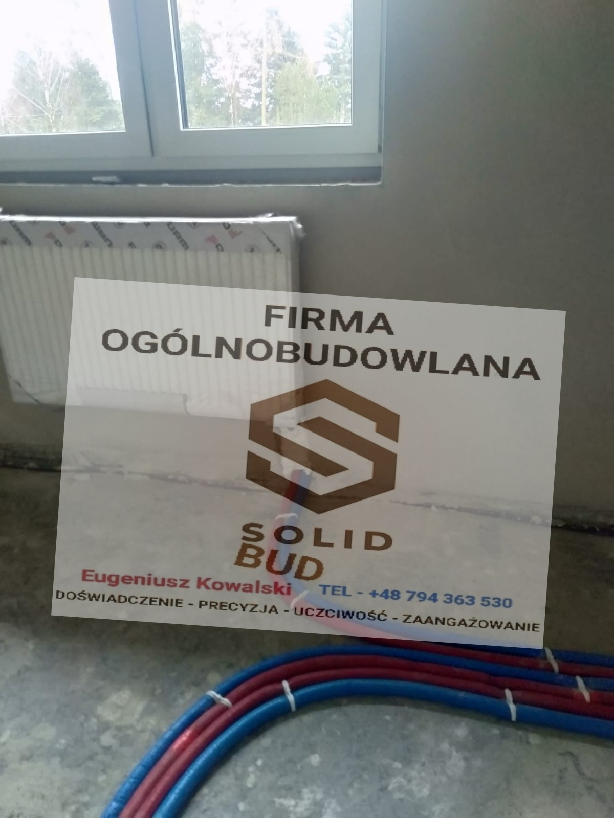 Tabliczka reklamowa firmy ogólnobudowlanej Solid Bud z logo, danymi kontaktowymi i hasłem reklamowym, ustawiona w surowym wnętrzu budowy z widocznymi rurami instalacyjnymi i grzejnikiem w folii.
