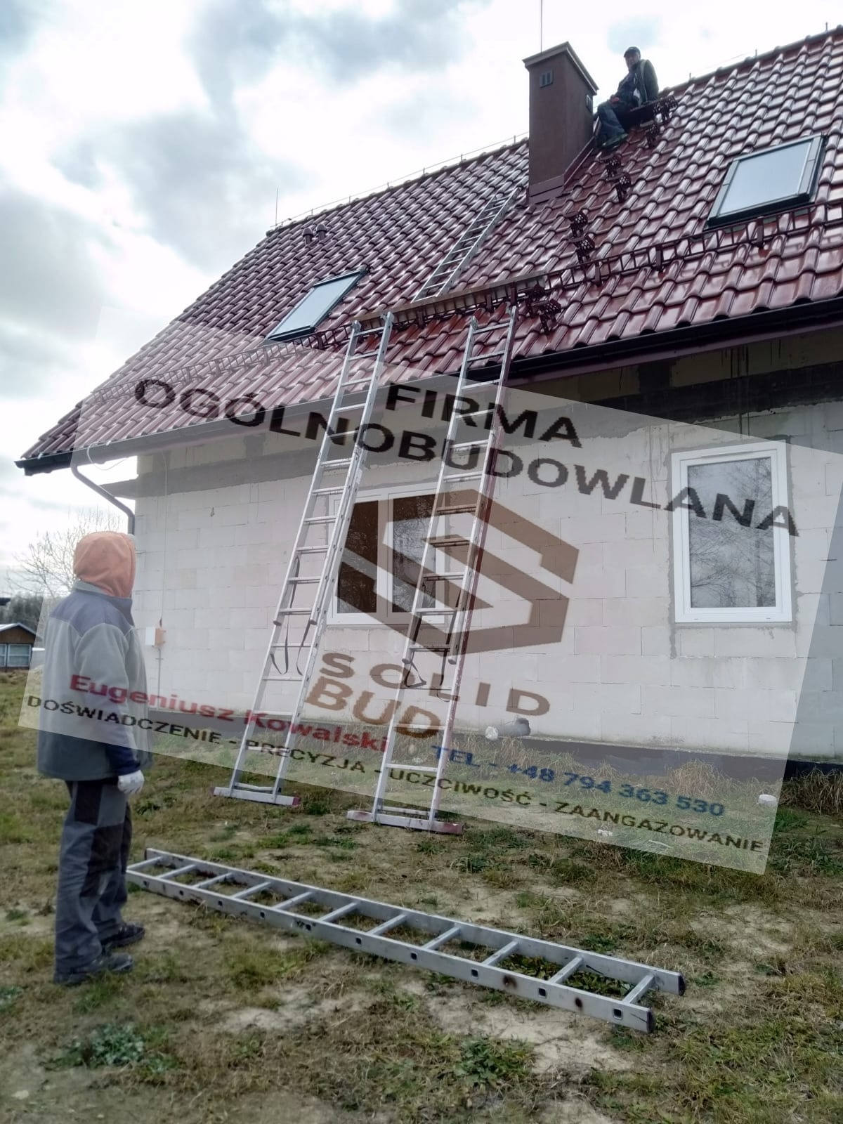 Pracownik w kombinezonie stoi obok domu z czerwonym dachem, na którym widać dwóch monterów, a do elewacji budynku przystawione są dwie aluminiowe drabiny.