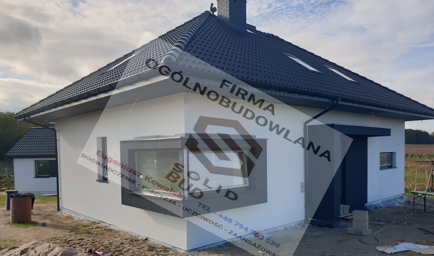 Biały dom jednorodzinny z ciemnoszarą stolarką okienną i dachem z dachówki ceramicznej, widoczny z boku, z elementami wykończeniowymi elewacji w trakcie prac, na pierwszym planie piasek i materiały...