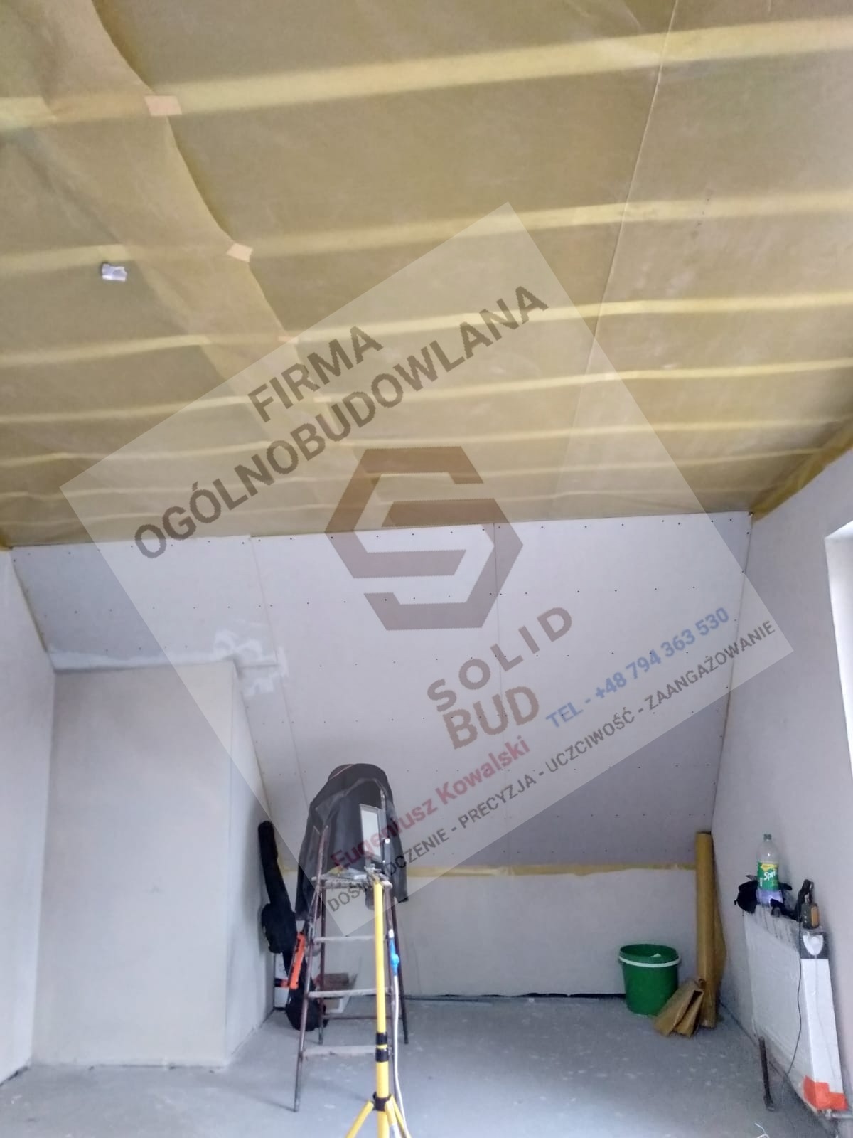 Ocieplone poddasze z widoczną izolacją i płytami gipsowo-kartonowymi, drabina i narzędzia budowlane w tle, logo firmy budowlanej na ścianie.