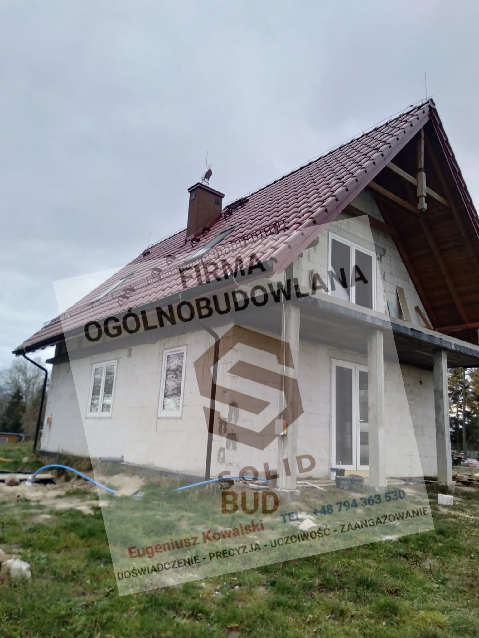 Dom w trakcie budowy, widoczne surowe ściany z bloczków, czerwona dachówka, niezakończone podbitki dachowe i fragment ogrodu z niebieskim wężem. Na zdjęciu widoczny jest także częściowo...