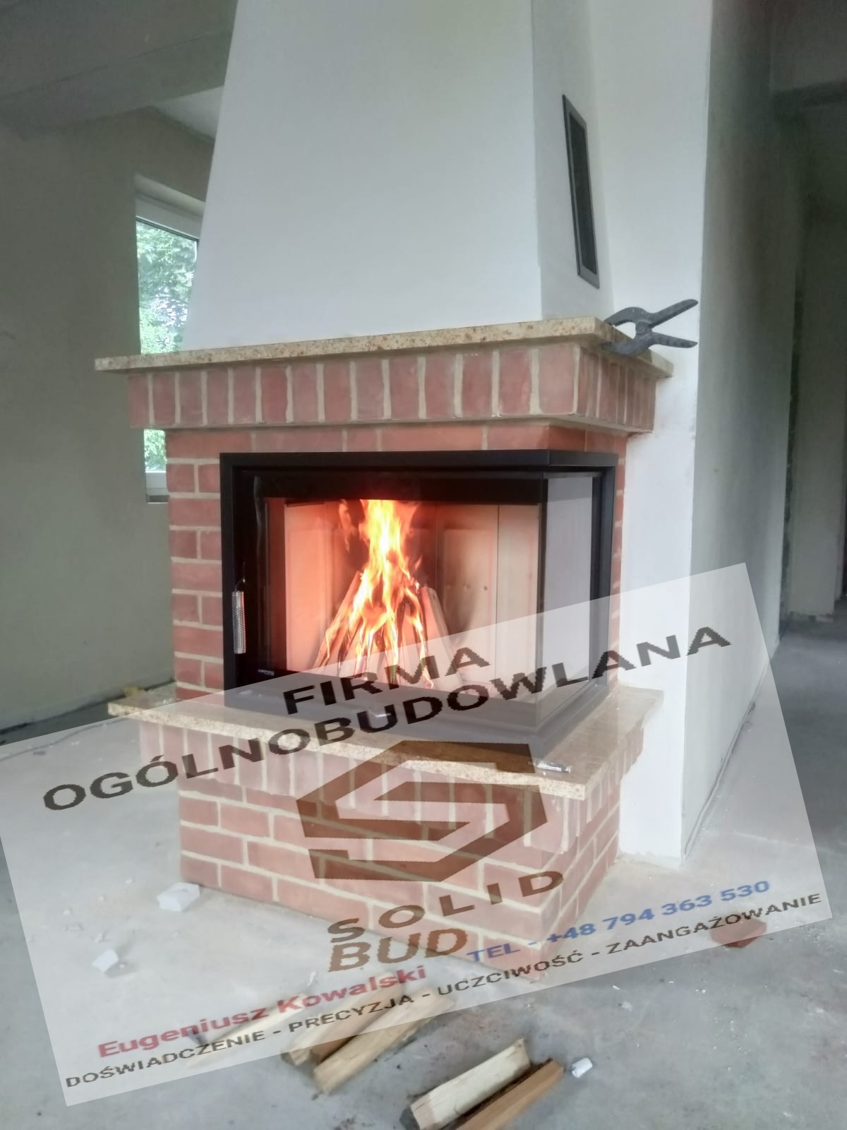 Murowany kominek z cegły klinkierowej z paleniskiem z płonącym drewnem, widoczne narzędzia kominkowe i logo firmy budowlanej na folii ochronnej.