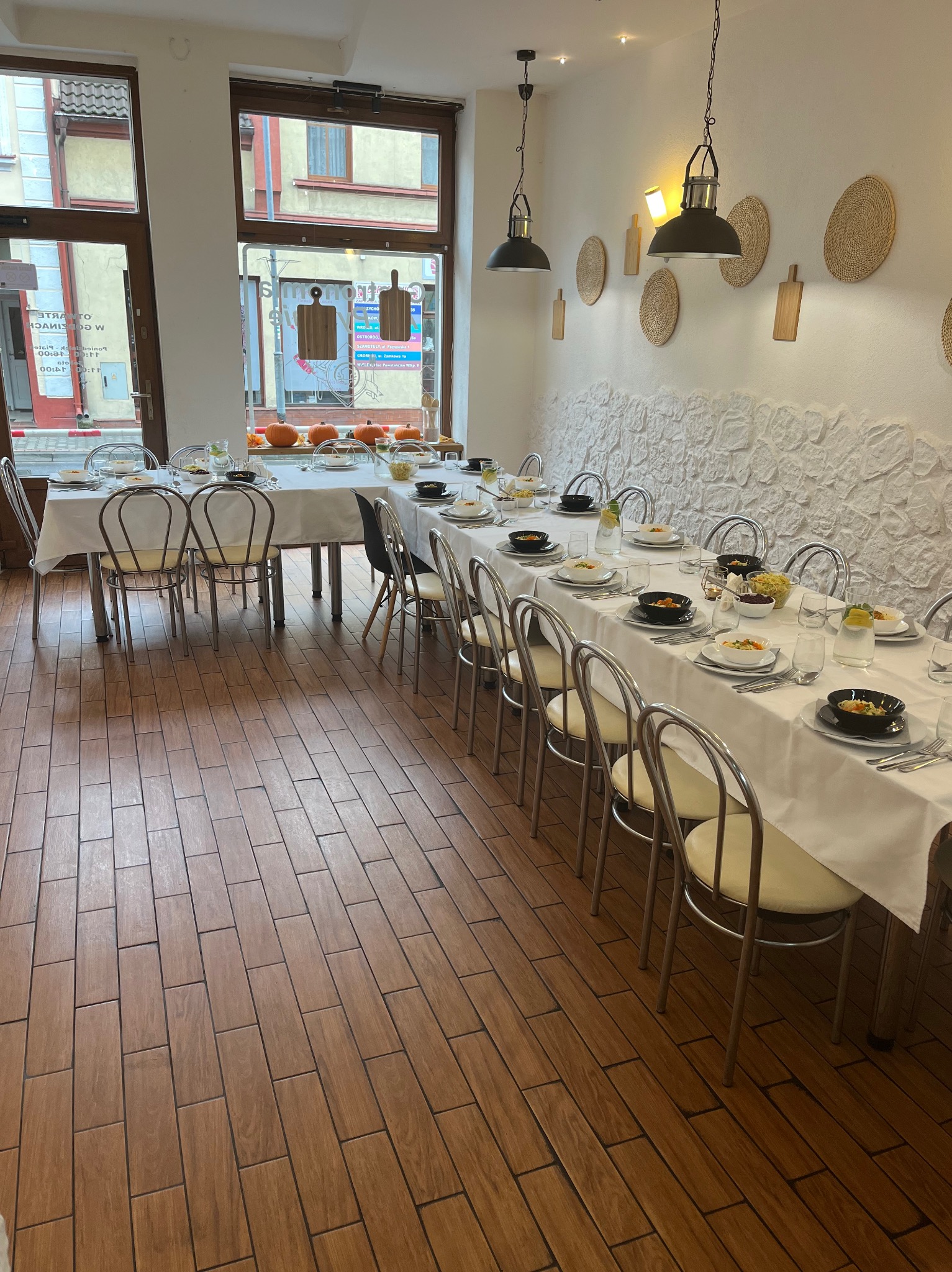Ustawiony stół z białym obrusem, nakryciami i dekoracjami w postaci dyń, w jasnym wnętrzu restauracji z drewnianą podłogą i ozdobami na ścianie.