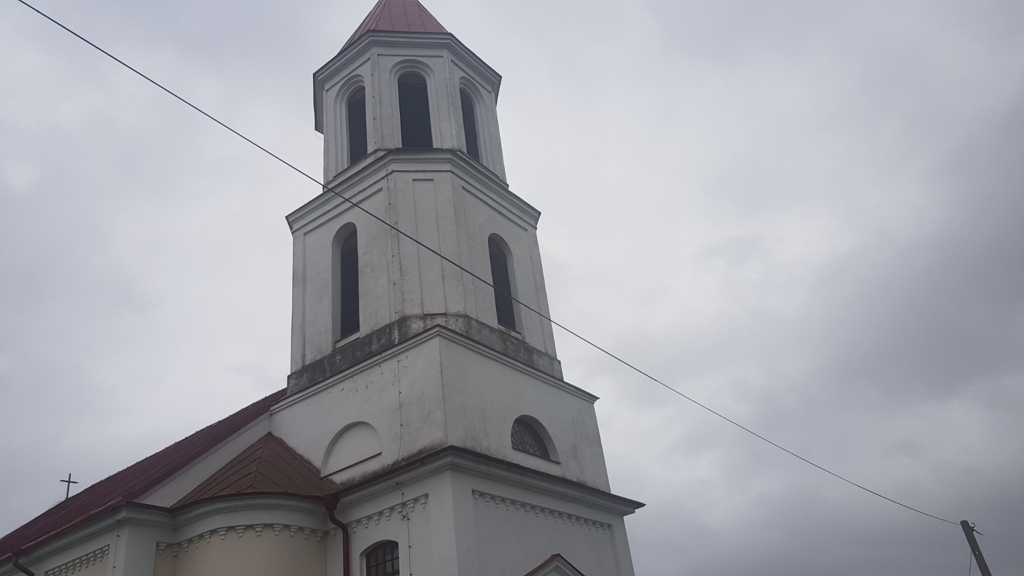 Biała wieża kościoła z czerwonym dachem na tle pochmurnego nieba, widoczne przewody elektryczne przecinające kadr.