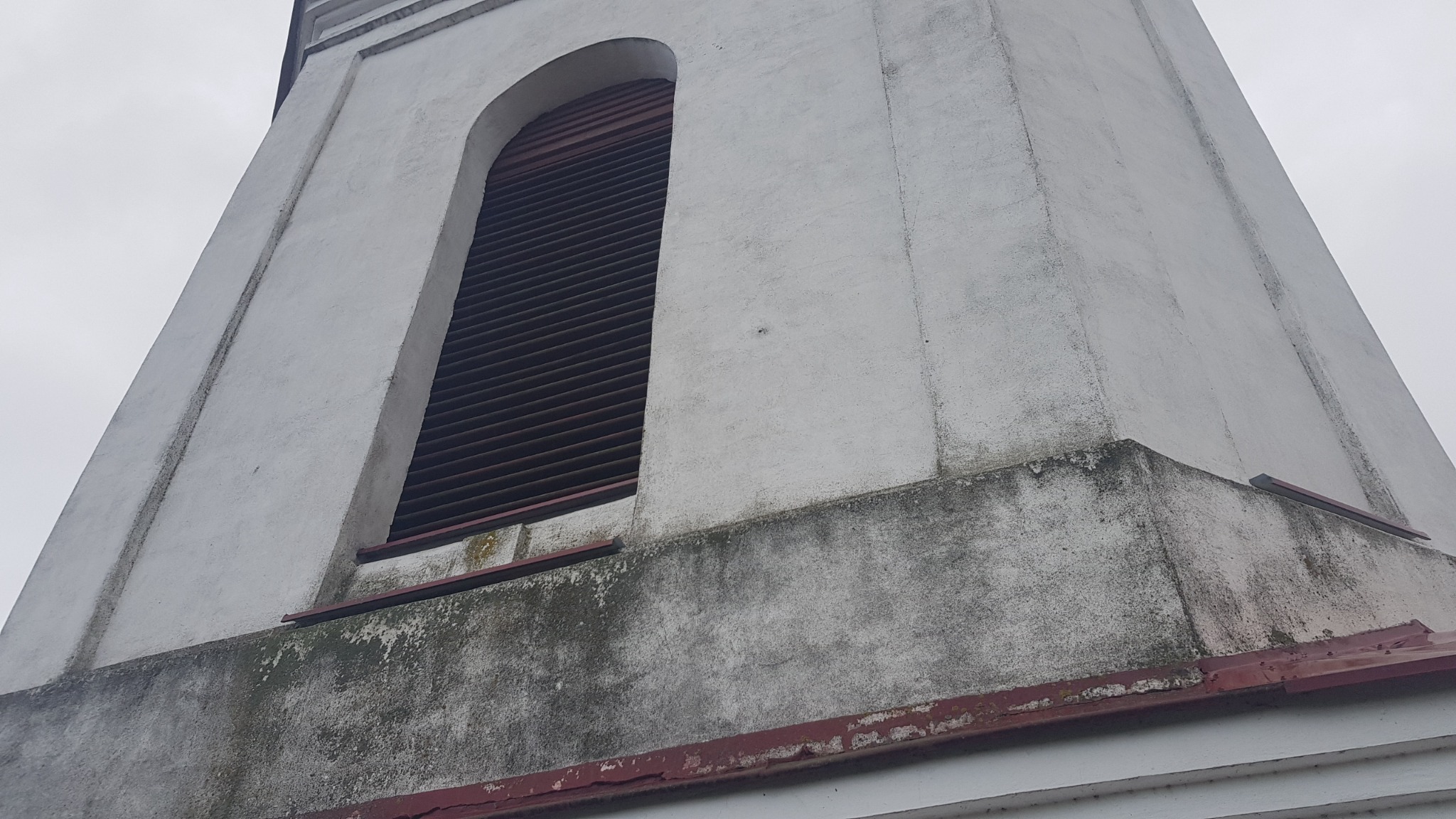 Fragment wieży kościelnej z wyraźnymi śladami wilgoci i zabrudzeń na szarej, szpachlowanej powierzchni, z widoczną kratką wentylacyjną w oknach.