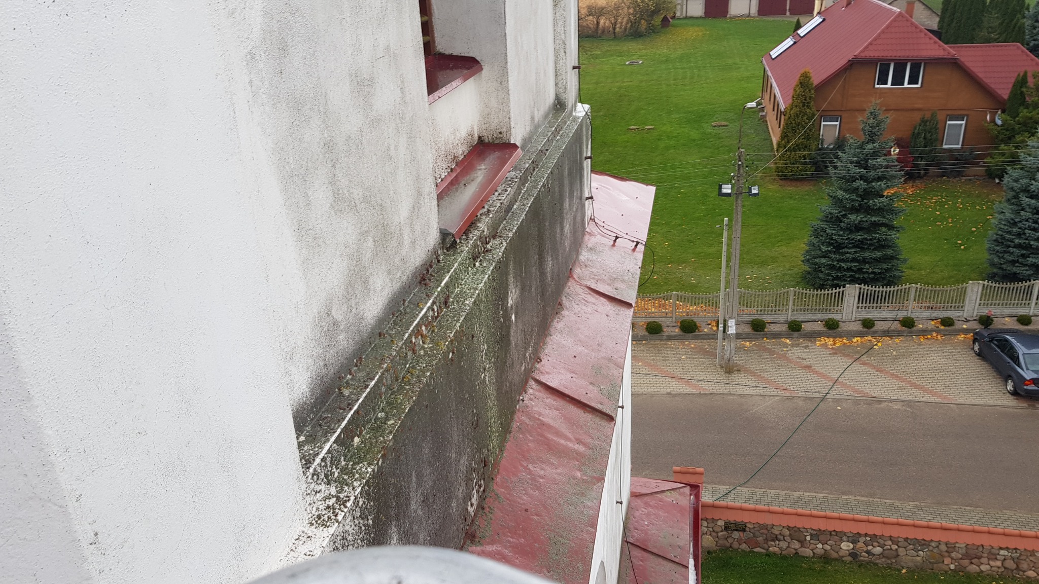 Brudna elewacja budynku z widocznymi śladami wilgoci i porostów, z perspektywy z góry, z czerwonymi elementami dachu i widokiem na zadbany ogród z domem w tle.