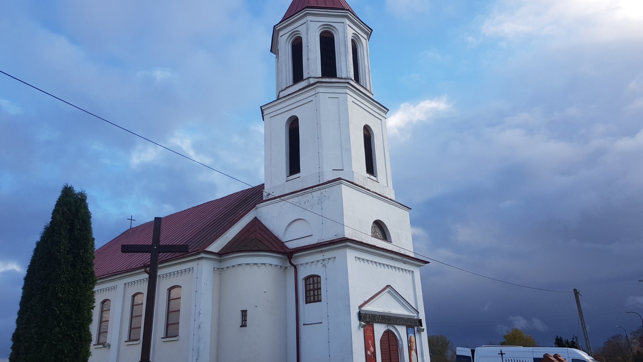 Biały kościół z czerwoną dachówką i wieżą na tle pochmurnego nieba, widoczny drewniany krzyż i wysoki cyprys.