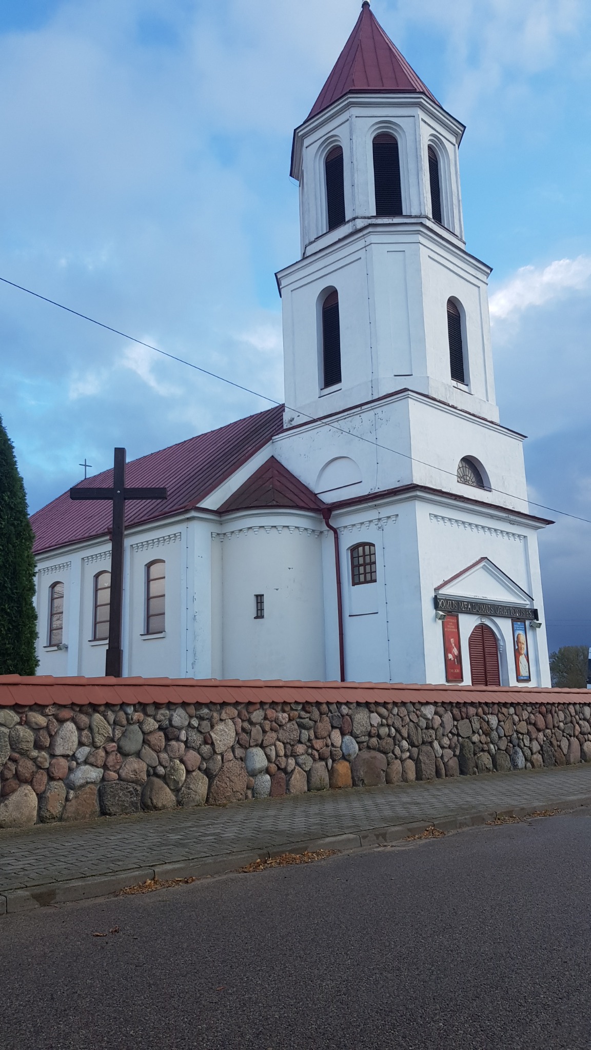 Biały kościół z czerwoną dachówką i wieżą, otoczony kamiennym murem z czerwoną dachówką, widziany z poziomu ulicy pod pochmurnym niebem.
