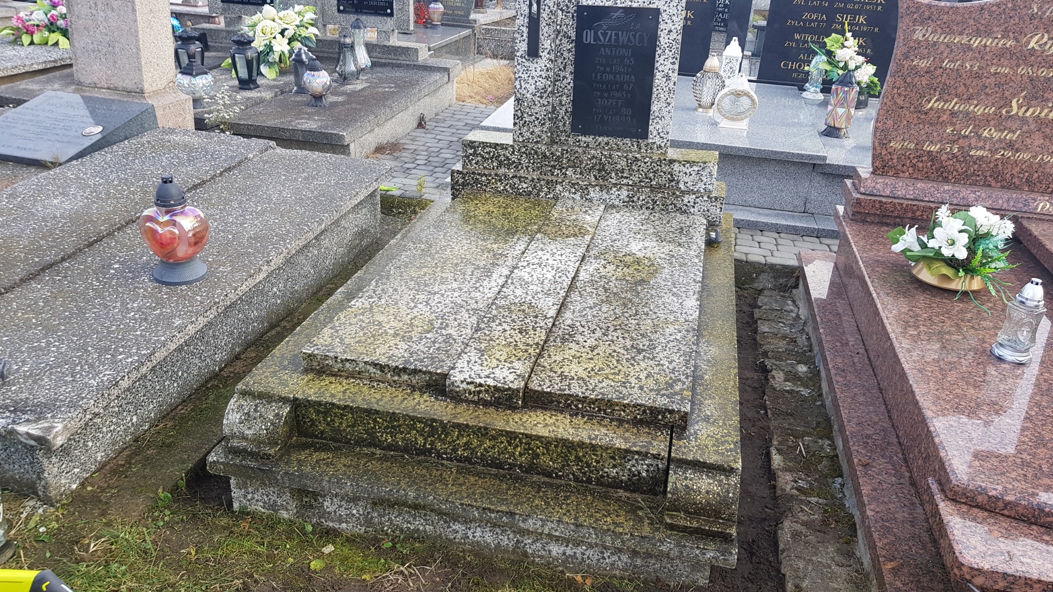 Zaniedbany, wilgotny nagrobek z szarego granitu, pokryty mchem i porostami, otoczony innymi grobami na cmentarzu.