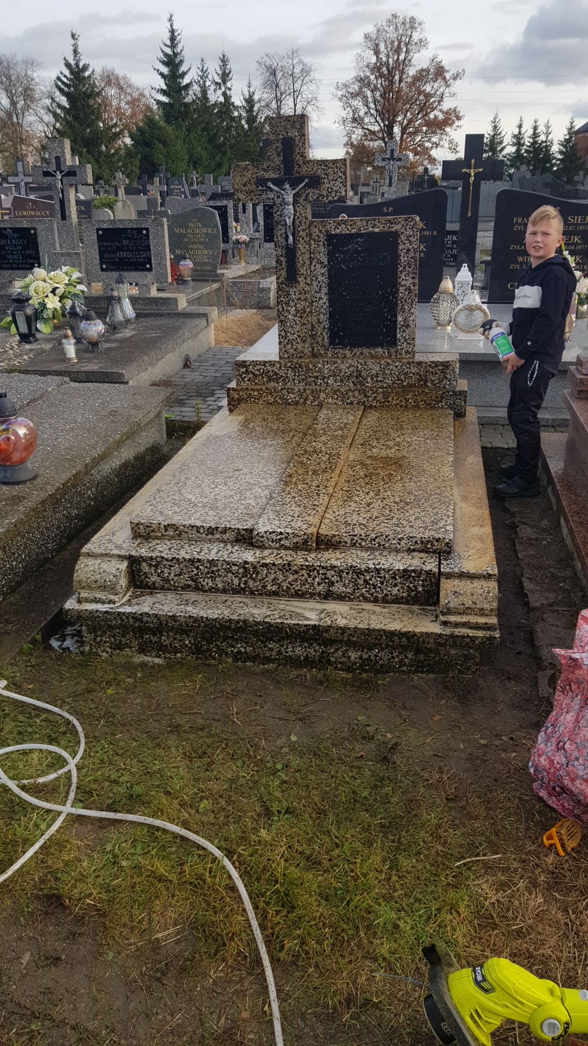 Chłopiec spryskuje płynem nagrobek z ciemnego kamienia na cmentarzu, widoczny krzyż i inne groby w tle, na ziemi leży biały wąż i żółta szorowarka.