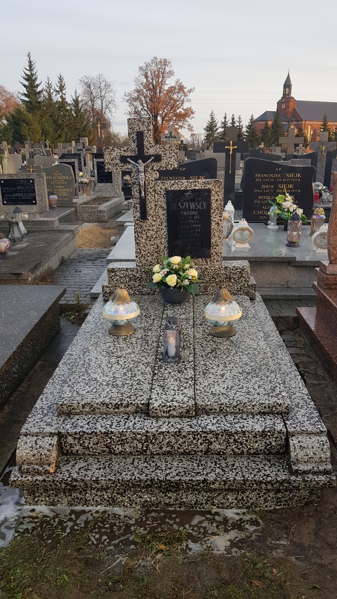 Granitowy nagrobek z krzyżem i tablicą inskrypcyjną, ozdobiony kwiatami w doniczce i dwoma lampionami z efektem tęczy, na tle cmentarza z innymi grobami i kościołem w oddali.