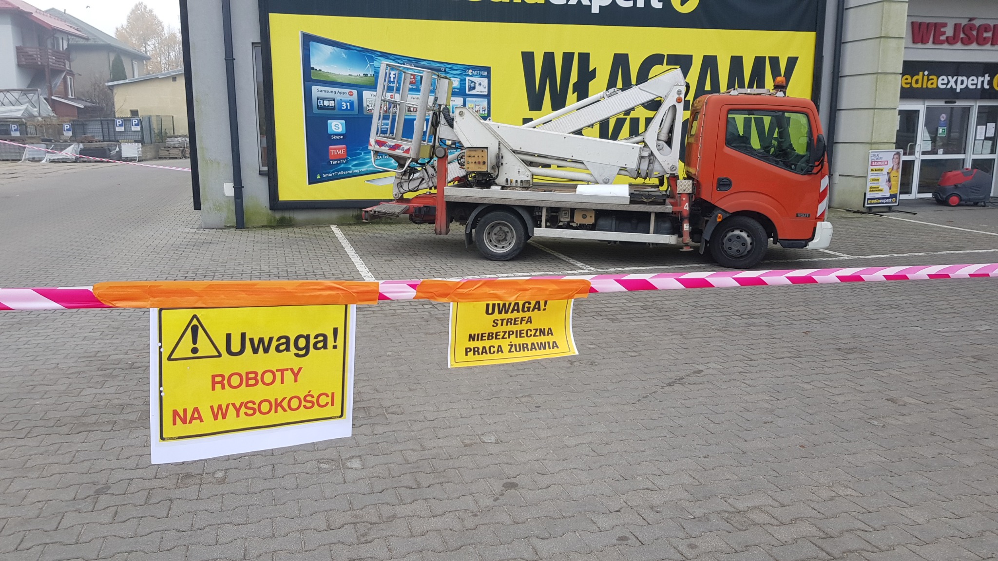 Pomarańczowy samochód z podnośnikiem koszowym zaparkowany przed budynkiem sklepu Media Expert, otoczony taśmą ostrzegawczą i znakami informującymi o pracach na wysokości i strefie niebezpiecznej...