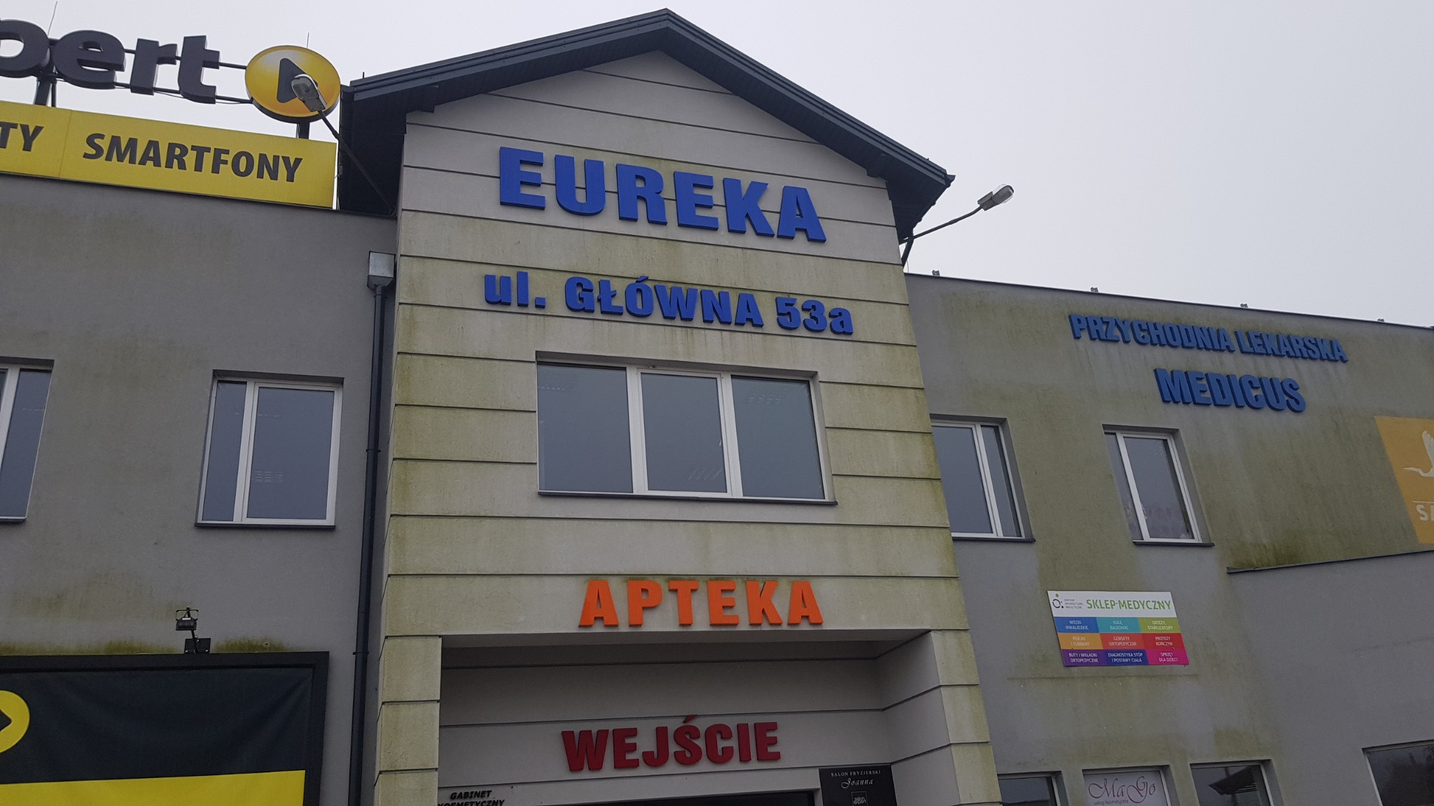 Budynek z wieloma szyldami: Expert Smartfony, Eureka ul. Główna 53a, Apteka, Wejście, Przychodnia Lekarska Medicus, Sklep Medyczny, Salon Fryzjerski Joanna, MaGo. Elewacja budynku jest lekko...