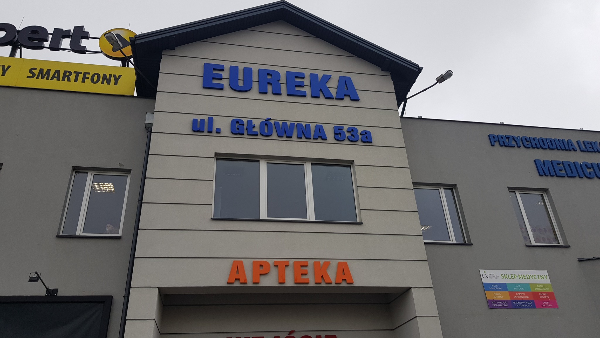 Fasada budynku z szyldami firm: Eureka, Apteka, Przychodnia Lekarska Medicus, Sklep Medyczny, oraz Expert Smartfony na górze. Widoczne okna z odbiciami w szybach i osoba w oknie po lewej.