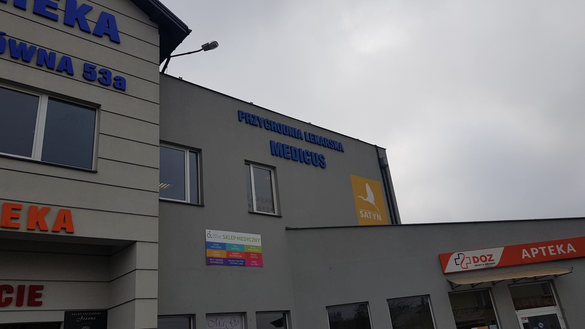 Elewacja budynku z napisem 'PRZYCHODNIA LEKARSKA MEDICUS' wykonanym z niebieskich liter, widoczny również szyld 'SATYN' z symbolem bociana, apteka oraz sklep medyczny.