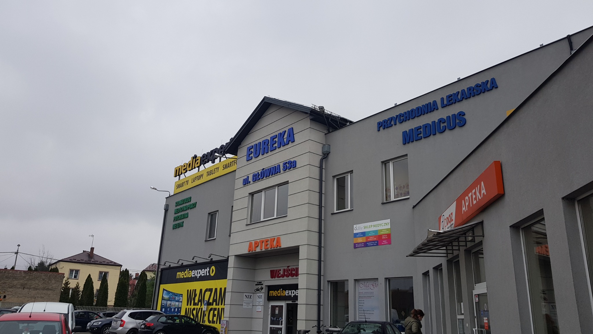 Szary budynek z niebieskimi napisami 'EUREKA' i 'PRZYCHODNIA LEKARSKA MEDICUS', z szyldami 'APTEKA' i 'mediaexpert' oraz widocznymi samochodami na parkingu w pochmurny dzień.