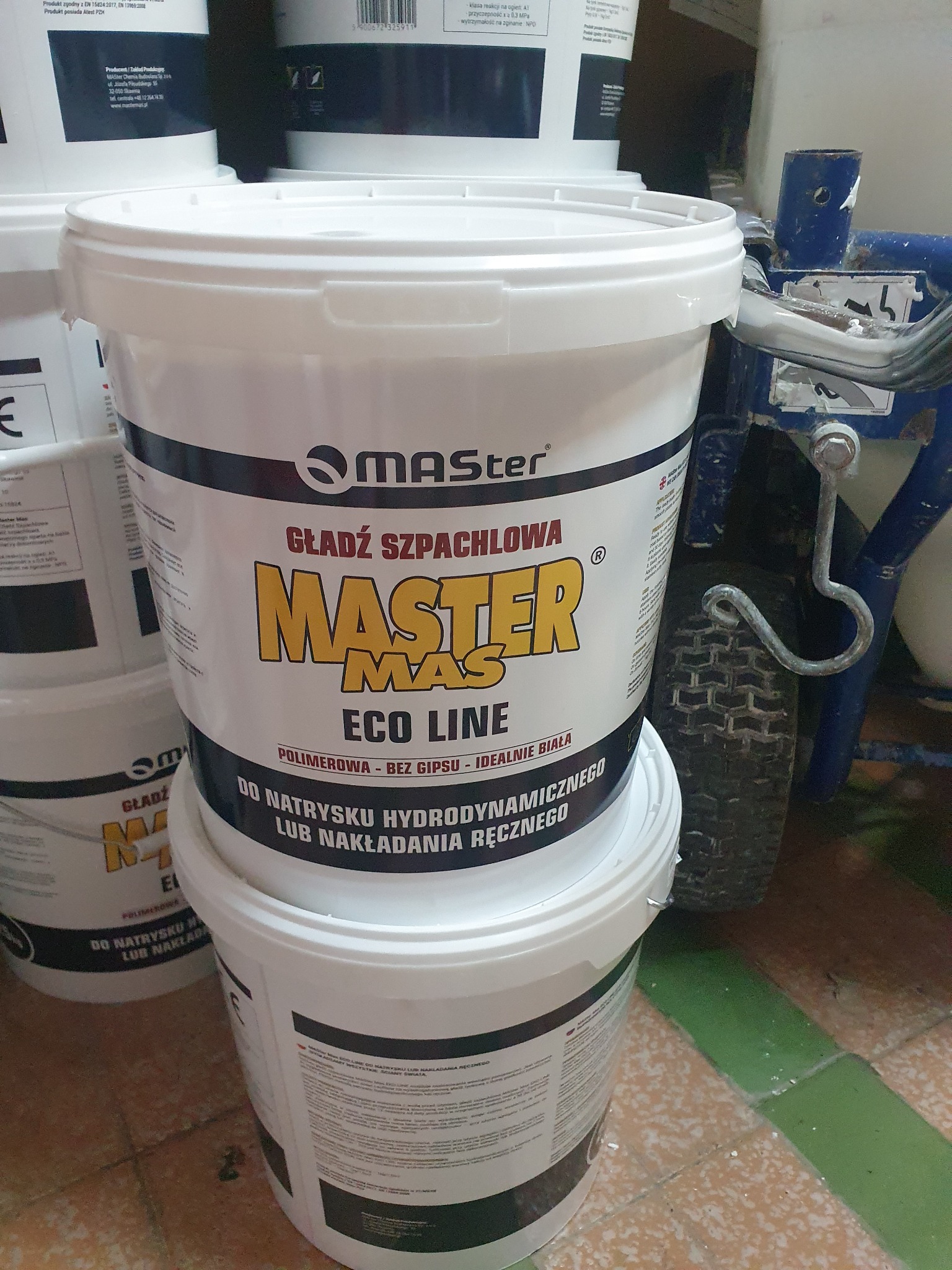 Białe wiadro z etykietą 'Gładź Szpachlowa Master Eco Line' ustawione na innym wiadrze, obok widoczny fragment urządzenia do natrysku hydrodynamicznego.