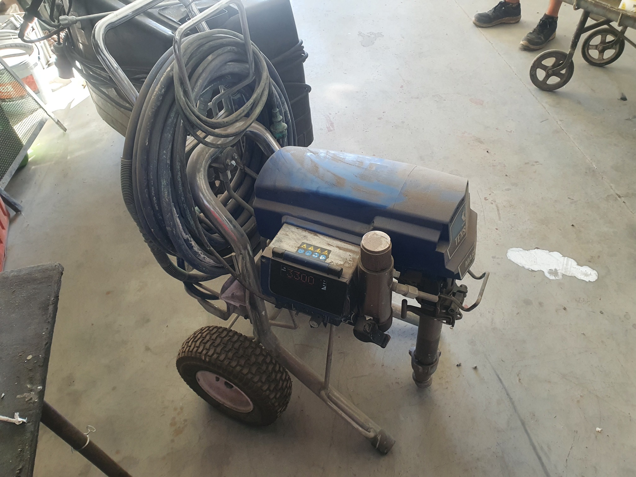 Profesjonalna, choć zakurzona, maszyna do natryskowego malowania z grubym wężem i oponą, stojąca na betonowej podłodze. W tle widoczne stopy osoby obsługującej urządzenie.