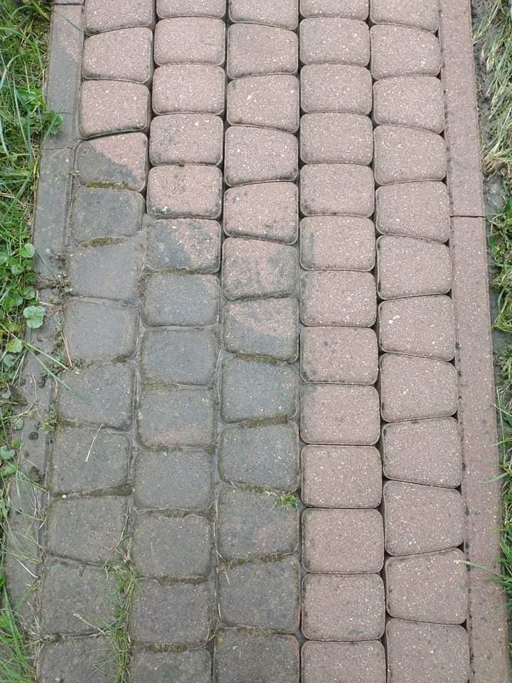 Częściowo oczyszczona kostka brukowa, widoczna różnica w kolorze i czystości między wyczyszczoną a zabrudzoną powierzchnią, zarośnięta trawą na obrzeżach.