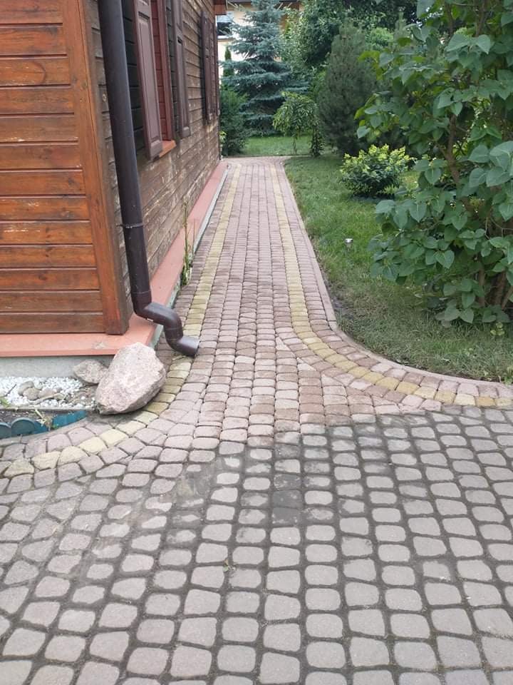 Ułożona kostka brukowa w odcieniach szarości i beżu prowadząca wzdłuż drewnianego domu z brązową rynną. Fragment kostki po prawej stronie jest mokry.