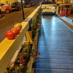 Sęk i Wiór - Miejska instalacja z drewna: podest z donicami z kwiatami, pomidory i dynia na barierce, w tle ulica z samochodami i budynki. Nietypowe zagospodarowanie przestrzeni.