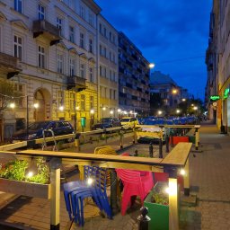 Sęk i Wiór - Oświetlony, drewniany ogródek kawiarniany z kwiatami i kolorowymi krzesłami na chodniku miejskiej ulicy nocą. W tle budynki i zaparkowane samochody.
