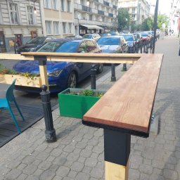 Sęk i Wiór - Uliczny bar z drewnianym blatem i donicami z kwiatami, ustawiony wzdłuż chodnika. W tle zaparkowane samochody i budynki miejskie. Widoczny przechodzień.