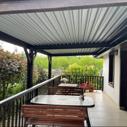 Sęk i Wiór - Zadaszony balkon z ciemnym drewnianym stelażem i jasnym, falistym dachem, wyposażony w dwa zestawy drewnianych mebli ogrodowych na beżowych płytkach, z widokiem na zielony krajobraz.