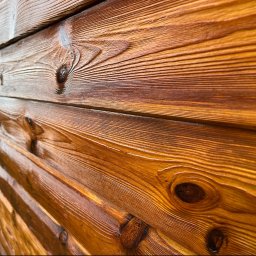 Sęk i Wiór - Blisko ujęte, lśniące drewniane deski z wyraźnymi słojami i sękami, podkreślające naturalną fakturę drewna po obróbce. Ciepły, brązowy odcień.