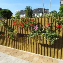 Ozdobny murek z prefabrykatów betonowych, z wnękami wypełnionymi kwitnącymi pelargoniami i innymi kwiatami, tworzący zieloną ścianę w ogrodzie.