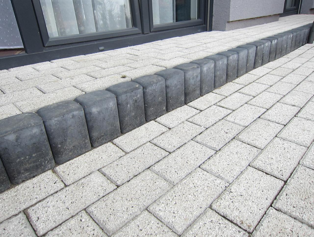 Jasnoszara kostka brukowa ułożona w jodełkę, obrzeża z ciemnoszarych kostek, widok z niskiej perspektywy przy ścianie budynku z oknem.